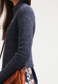 Pull en tricot bleu marine avec un col montant, une texture côtelée et un design court, associé à un sac en cuir marron et une jupe fleurie.