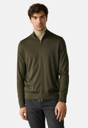 Mann trägt dunkelgrünen Zip-up-Pullover über beige Rollkragenpullover und dunkelgraue Jeans, steht vor einfachem hellem Hintergrund.