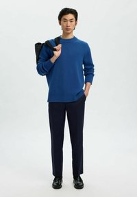 Hombre con suéter azul y pantalones oscuros, llevando una bolsa negra al hombro, de pie frente a un fondo blanco.