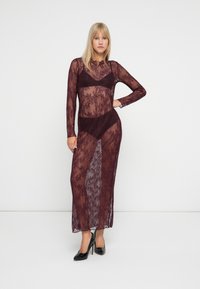 Blondīne, kas ģērbusies garā, caurspīdīgā bordo mežģīņu kleitā virs atbilstoša krūštura un apakšveļas, apavi ar melnām augstiem papēžiem, stāv pret baltu sienu.