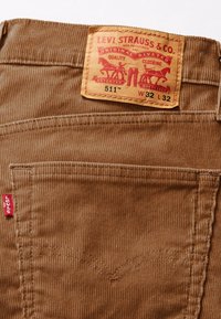 Brązowa tkanina sztruksowa z wąskimi pionowymi żebrowaniami, metka Levi's z czerwonymi akcentami, dodatkowe kieszenie i klasyczne detale szycia.