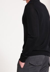 Svart stickad tröja med rund halsringning, långa ärmar och ribbade manschetter, i kombination med grå byxor med klafffickor och en knappdetalj.