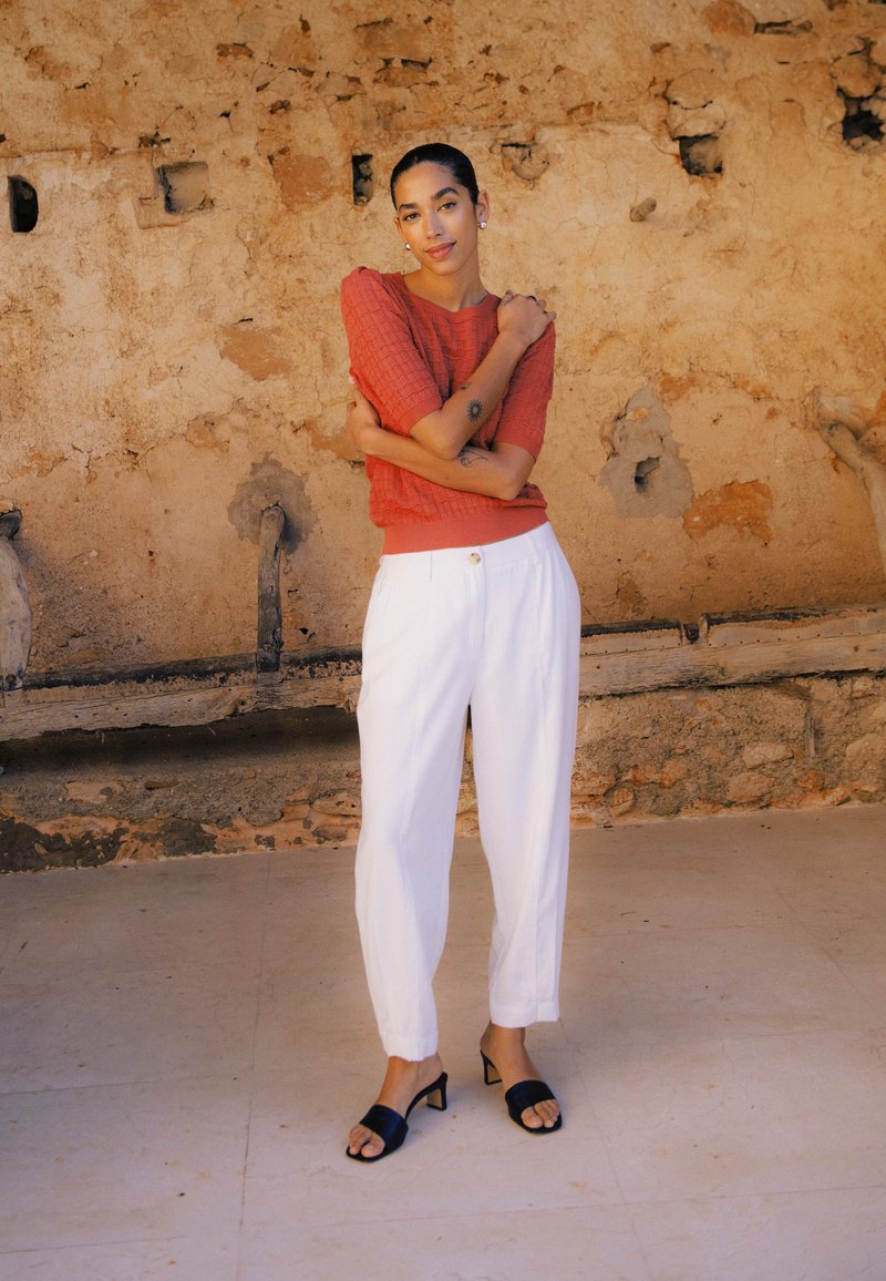 Frau mit kurzen dunklen Haaren in einem orangefarbenen Strickoberteil, weißen Hosen und schwarzen Absätzen steht mit verschränkten Armen vor einer rustikalen, strukturierten Wand.