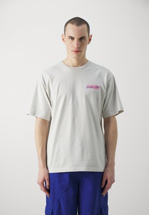 Camiseta de color beige claro con mangas cortas, con texto bordado en rosa en el lado izquierdo del pecho. Material de algodón suave, corte relajado.