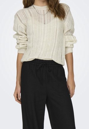 Femme portant un pull en maille côtelée crème avec des manches bouffantes et un pantalon noir à cordon de serrage, sur un fond uni.