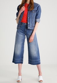 Veste en denim, bleu foncé avec des manches retroussées; top en dentelle rouge; jeans larges, bleu clair avec un ourlet effiloché; chaussures blanches à bout ouvert.
