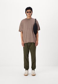 Barna oversize póló, olívazöld, tapered nadrág rugalmas szárakkal, fehér sportcipő barna talppal, fekete keresztpántos táska, kerek napszemüveg.