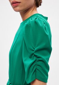 Πράσινο φόρεμα με εφαρμοστό σιλούετ, υφασμάτινα φουσκωτά μα�νίκια και λεία υφή. Η πλευρική άποψη αναδεικνύει λεπτομέρειες των μανικιών και της λαιμόκοψης.