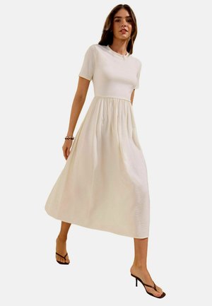 Donna che cammina, indossa un vestito midi color crema a maniche corte con vita raccolta e sandali neri con tacco.