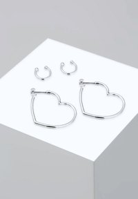 Pendientes de aro plateados en forma de corazón; superficie lisa y brillante. Incluye dos pendientes circulares más pequeños. Presentados en un pedestal claro.