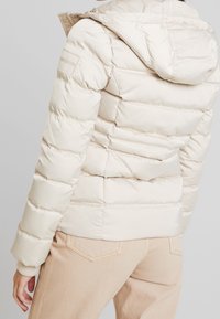 Giacca puffer crema con cuciture orizzontali, cappuccio aderente e polsini a coste, realizzata in materiale liscio e lucido. Presenta una toppa con logo.