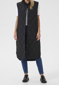 Czarna pikowana bezrękawnik z wysokim kołnierzem, zatrzaskami z przodu i długim krojem, zestawiony z niebieskimi dżinsami i czarnymi butami.