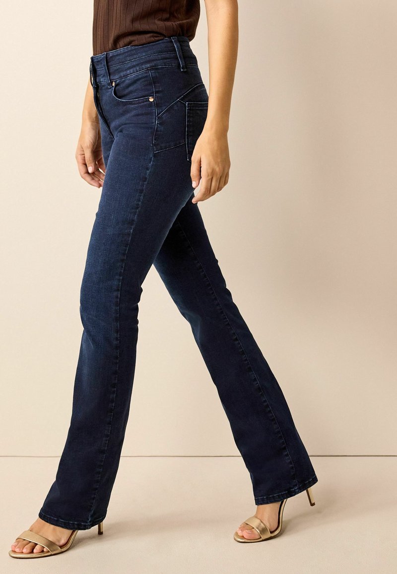 Jeans de mezclilla de color azul oscuro con campana y cintura alta, con detalles de costura y cremallera frontal. Combinados con sandalias de tacón dorado.