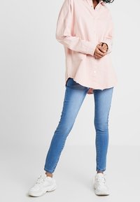 Ljusrosa överdimensionerad knapp-up skjorta med rundad nederkant, parat med åtsittande blå jeans och vita sneakers.