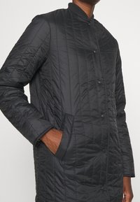 Chaqueta acolchada negra con cuello alzado, botones de presión, dos bolsillos laterales y puños acanalados. Textura suave y diseño ligero.