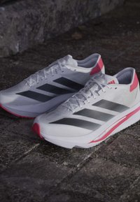 Hvide Adidas løbesko med sorte striber og pink detaljer på en stenbelagt overflade.