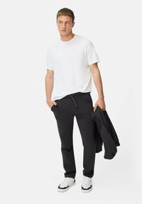 Witte t-shirt met korte mouwen, zwarte joggingsbroek met zakken en trekkoord, en witte sneakers met blauwe accenten, vastgehouden met een zwarte hoodie.