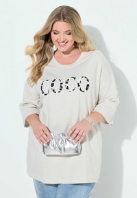 Lysebeige oversized t-shirt med "COCO" i sorte og brune leopardprintbogstaver, kombineret med en metallic sølv clutch og denim.