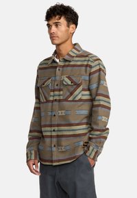 Camicia a quadri marrone con strisce blu e verdi, due tasche sul petto, chiusura con bottoni e tessuto textured. Etichetta nera con logo sull'orlo.