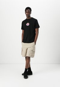 Svart bomull T-skjorte med et fargerikt grafisk print på forsiden, kombinert med beige cargo-shorts med flere lommer og en avslappet passform. Sorte joggesko.