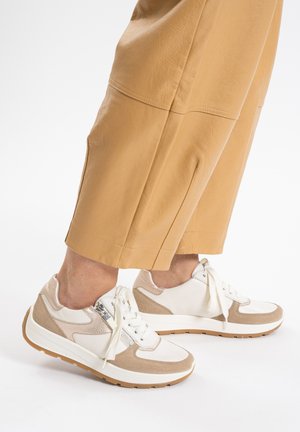 White and beige sneakers with suede and leather panels, rounded shape, flat laces, and a textured sole. Worn with tan wide-legged pants.
