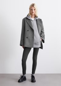 Cappotto grigio doppiopetto sopra una felpa grigia con cappuccio, abbinato a leggings scuri e scarpe nere con lacci. Texture morbide, vestibilità rilassata, dettagli minimi.