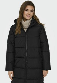 Svart dunjakke med glidelås foran, horisontale sømmer og hette. Lange ermer og sidelommer. Bært over en lys beige turtleneck.