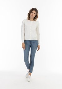 Cardigan bianco a maniche lunghe con chiusura a bottoni, abbinato a jeans blu e sneaker bianche. Tessuto morbido, vestibilità casual.