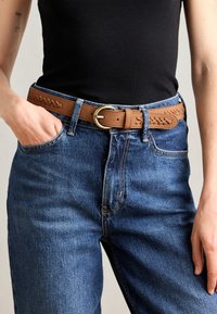 Brown leather belt with woven detailing, gold-toned circular buckle, worn over blue denim jeans, with a black tank top visible.