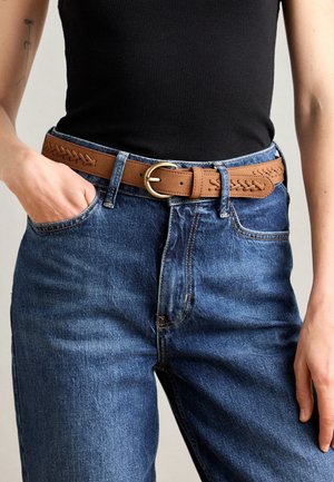 Brun læderbælte med vævet detalje, guldtonet cirkulær spænde, båret over blå denimjeans, med en sort tanktop synlig.