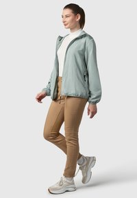 Gaiši zaļa jaka ar rāvējslēdzēja aizdari, elastīgiem aprocēm un divām sānu kabatām, kombinēta ar pieguļošām bēša krāsas biksēm un bēša sporta apaviem.