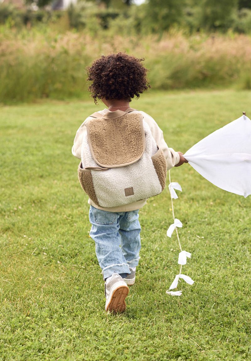 Un sac à dos tan et crème avec une texture duveteuse et un rabat arrondi, porté par un enfant tenant un drapeau blanc, marchant sur de l'herbe.