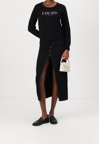 Femme portant un haut à manches longues noir avec le texte "LIU·JO 1995", une jupe noire fendue à l'avant, des mocassins noirs, tenant un petit sac à main blanc.