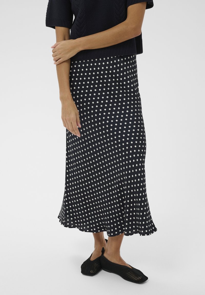 Kobieta ubrana w czarną spódnicę midi w białe kropki, zestawioną z czarnym dzianinowym topem i czarnymi siateczkowymi butami wsuwanymi.