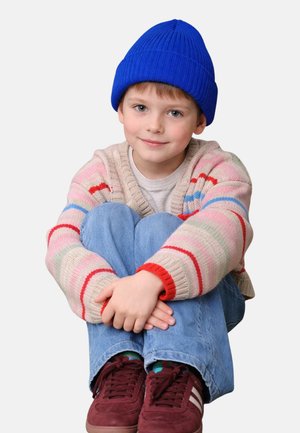 Knitted sweater with pastel stripes, blue beanie, gray t-shirt, denim jeans, and maroon sneakers. Child sitting with arms wrapped around knees.