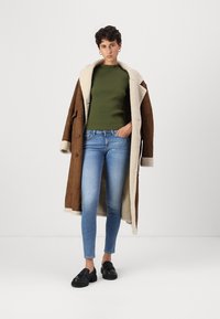 Bruin shearling-voering jas, groene geribbelde trui, blauwe skinny jeans en zwarte instapschoenen. De jas heeft grote knopen en een gestructureerde stof.