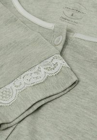 Šedá texturovaná tkanina s bílou krajkovou ozdobou na rukávu, se zapínáním na knoflík a kulatým výstřihem. Logo "Intimissimi" viditelné na tričku.