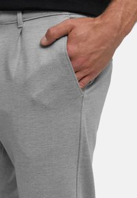 Miehen oikea käsi työnnettynä vaaleanharmaiden housujen etutaskuun neutraalia taustaa vasten.