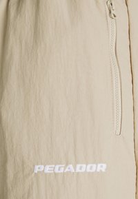 Béžové lehké šortky s bočním zipem, hladkým povrchem a vyšitým logem "PEGADOR" v bílé barvě na spodním lemu.
