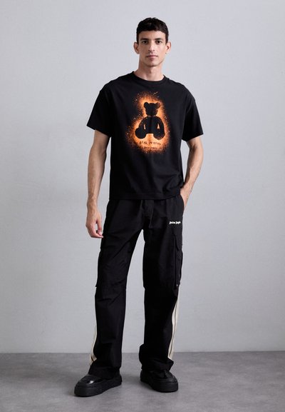 Black t-shirt with orange graphic of a bear and text, paired with black cargo pants featuring side stripes and logo. Footwear is black platform sneakers.