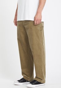 Pantalons en velours côtelé kaki avec une coupe droite, dotés d'une poche latérale et de la cheville du porteur visible dans des baskets noires.