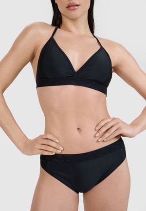 Kobieta ubrana w czarny dwuczęściowy bikini z cienkimi ramiączkami i szerokim paskiem, pozująca z rękami na biodrach na jednolitym tle.