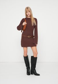 Pruun soonikkoeline kootud kleit, pikkade varrukatega, taljest pruun vööga seotult. Kombineeritud mustade põlveni ulatuvate saapade ja pruuni kotiga.