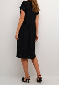 Vestido midi negro con mangas cortas, patrón de ojales texturizado y costura en la cintura. Combinado con sandalias de tacón negras, mostrando un acabado de tela suave.