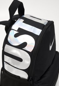 Fekete hátizsák fényes, oversized, fényvisszaverő "JUST DO IT" logóval, textur�ált anyaggal és cipzáras rekeszekkel. A Nike logó látható.