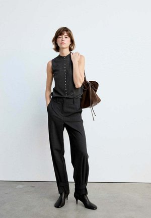 Femme portant un haut noir sans manches boutonné, un pantalon noir taille haute, des bottines noires et un sac à main en cuir marron porté à l'épaule.