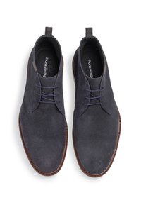 Zapatillas de lona azul marino con una superficie texturizada, con acentos marrones, diseño de cordones y una pestaña en el talón. Suela plana.