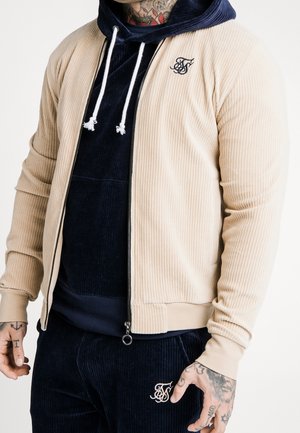 Homme portant une veste côtelée beige zippée par-dessus un sweat à capuche bleu marine et un pantalon assorti avec des tatouages visibles sur les mains et le cou.