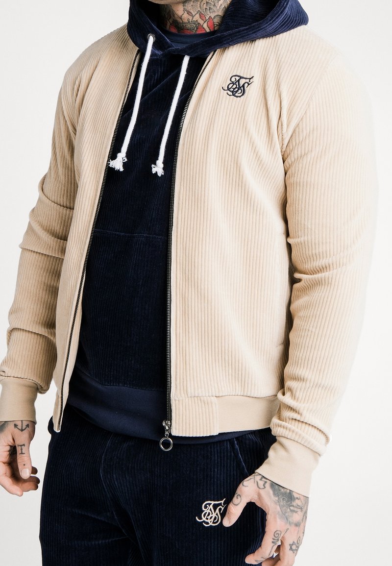 Homme portant une veste côtelée beige zippée par-dessus un sweat à capuche bleu marine et un pantalon assorti avec des tatouages visibles sur les mains et le cou.