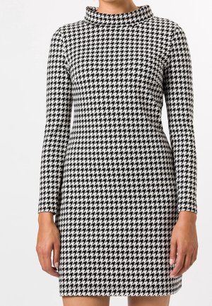Persona con un vestido de manga larga con estampado de pata de gallo en blanco y negro y cuello alto, de pie frente a un fondo liso.